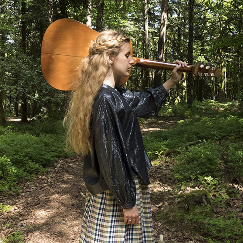 Femme de profil dans la forêt, guitare sur l'épaule, portant la chemise Billie en lurex noir, musique folk